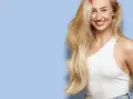 En leende kvinna med långt, blont hår som ser ut att bleka håret naturligt. Hon bär en vit topp och jeans mot en ljusblå bakgrund.