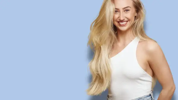 En leende kvinna med långt, blont hår som ser ut att bleka håret naturligt. Hon bär en vit topp och jeans mot en ljusblå bakgrund.